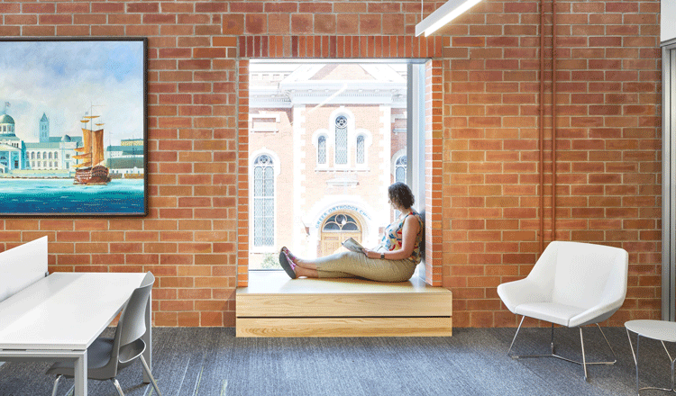 A woman sitting in a window seat inside the city of Kingston's Frontenac Central Branch Public Library. The wall is exposed brick with a work of art blue in hue showing a ship sailing in the ocean. There is a white table to the left of the window and a white chair to the right of the window.