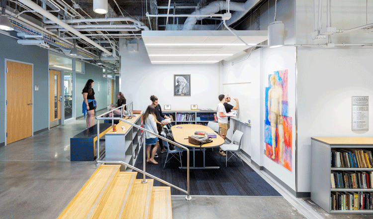 A photo of the Rita Hollings Science Center at the College of Charleston. The image shows four wooden steps leading to a cooridor wrapping around a sunken collaboration space with students working in a group and writing on a white board. The collaboration space contains half-stack bookshelves and colorful art deocating the wall.  