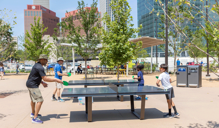 Four people playing ping pong at the Gene Leahy Mall with trees and city skyline of Omaha, Nebraska in the background.