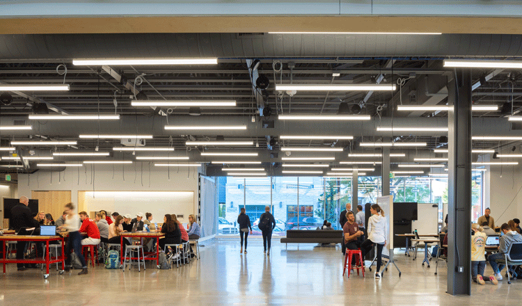 This photo shows students doing various activities in the University of Nebraska Johnny Carson Center for Emerging Media Arts. The room is large room with high ceilings and exposed HVAC equipment overhead. On the left, 10-15 students are listening to a lecture. In the middle, two people walk toward tall windows in the back of the space. On the right, 2-3 groups of 3-5 students use movable white boards on wheels to collaborate. 