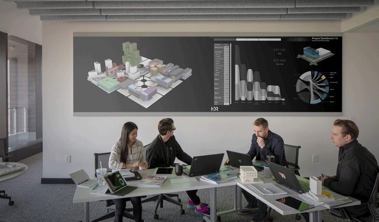 Data Driven Design team collaborating at tables with dashboard of software on the large screen behind them.