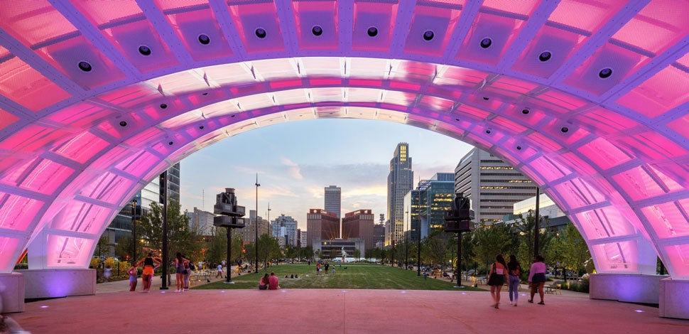 Bandshell in a park lit up with pink lights, with a city skyline in the background