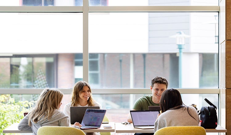 Students working together with computers at a table, with floor-to-ceiling windows behind them