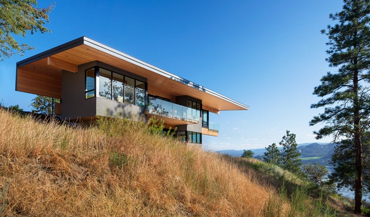 Mass timber house on a grassy hill, trees surrounding, blue sky