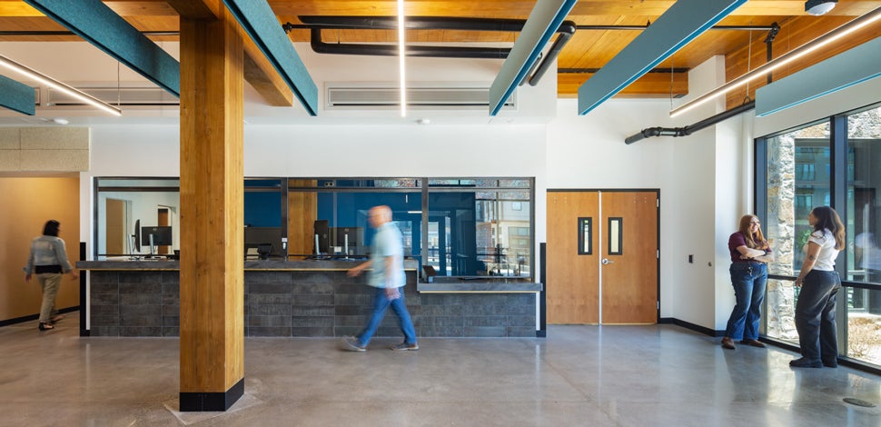 Ticket lobby of a bus station, wooden pillars, two people in the corner, person walking by