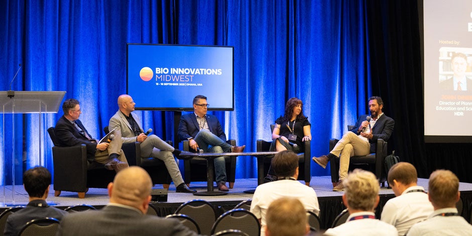 A panel of people sitting on a stage with a TV screen behind them