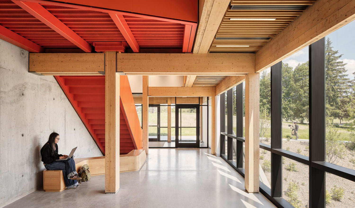 A building corridor with red detailing and timber interiors. A person sits with a computer on a bench